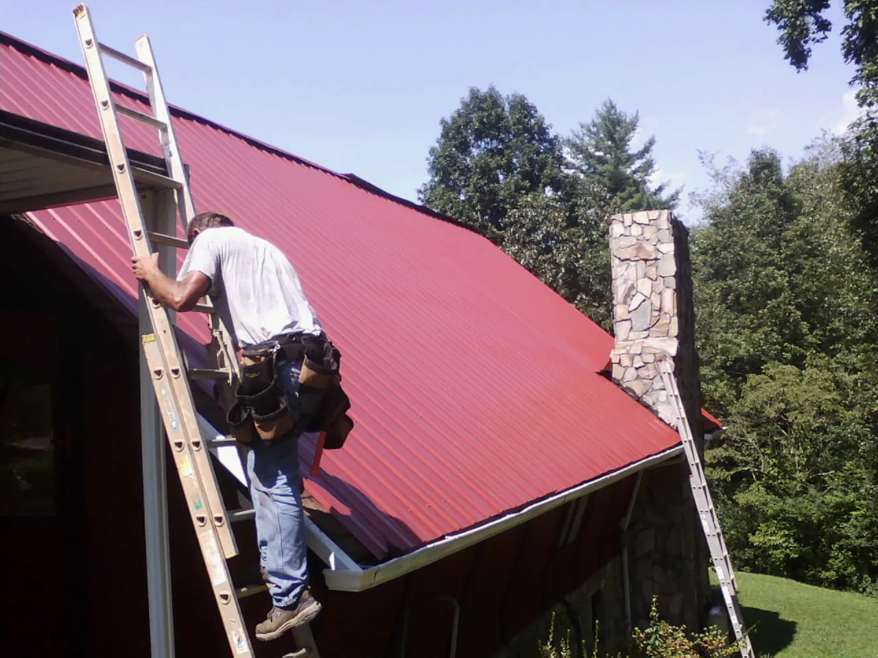 Roofer on a ladder beside a red metal roof and stone chimney — Soffit & Fascia Repair by Roofing Brothers in Nanuet